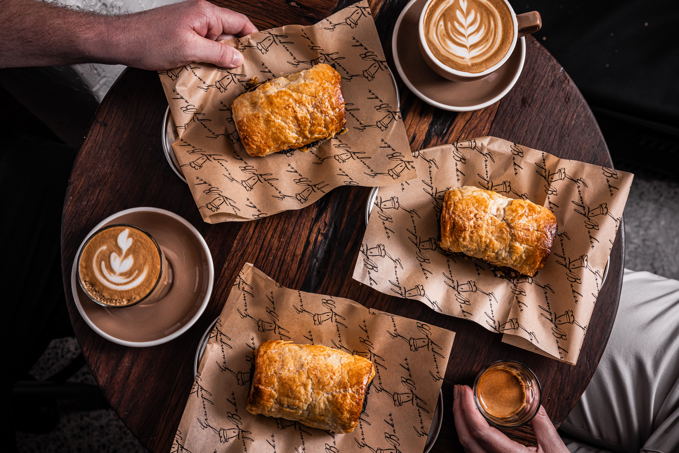 Sausage rolls on cafe table with coffees alongside two people dining